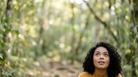 Young woman with curly dark hair and medium brown skin tone gazing upward in a lush forest setting. Suitable for wellness, nature, inspiration, and lifestyle marketing campaigns.