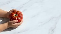 Hands holding fresh red ripe tomato on white marble background, organic produce for cooking, healthy food concept.
