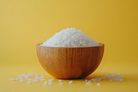 White rice grains in wooden bamboo bowl against vibrant yellow background, with scattered rice around base for food photography.