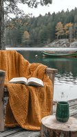 Cozy reading spot on wooden dock with mustard throw blanket, open book, and ceramic mug overlooking serene lake with canoe and autumn forest background.