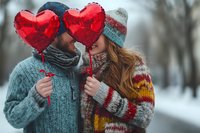 Young couple in winter clothing holding red heart balloons share romantic moment in snowy park setting, wearing cozy knit sweaters and warm accessories.