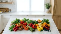 Fresh organic vegetables and fruits arranged on white marble kitchen counter including tomatoes, asparagus, leafy greens and colorful produce for healthy cooking.