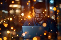 Young bearded man in glasses working on laptop at night with glowing bokeh lights and magical sparkle overlay effect creating cozy atmosphere.