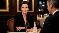 Elegant mature businesswoman in black suit enjoying wine dinner conversation with male colleague in upscale restaurant setting with golden frames.