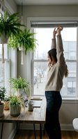 Young woman stretching arms up in cozy home office with plants by window. Remote work wellness and healthy lifestyle concept for productivity.