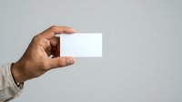 Male hand with medium skin tone holding blank white business card against neutral gray background for branding and corporate identity mockups.