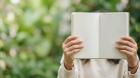 Open book held by person with hands visible, blank pages ready for content, outdoor green blurred background, education and reading concept for learning materials.