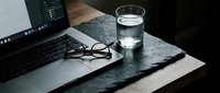 Laptop with glasses and glass of water on dark slate surface, moody workspace setup for productivity and remote work concept.