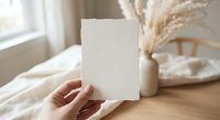 Hand holding blank deckle edge paper card in cozy boho interior with pampas grass, mockup for wedding invitation or stationery design.