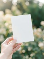 Hand holding blank white paper card against blurred green nature background with bokeh lights for mockup design and text placement.