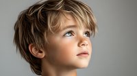 Young Caucasian child with tousled blonde hair and blue eyes gazing upward with wonder against neutral gray background, capturing innocence and curiosity.