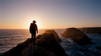 Silhouetted male traveler standing on rocky coastal cliff at sunset, gazing toward ocean horizon with golden sky and distant rock formations, adventure and exploration concept.