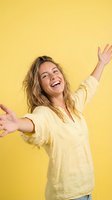 Happy woman with long brown hair and fair complexion wearing pale yellow linen shirt with arms outstretched against bright yellow background, expressing joy and freedom for lifestyle and wellness content.