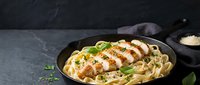 Grilled chicken breast sliced over creamy pasta with herbs in cast iron skillet on dark slate background for restaurant menu and food photography.