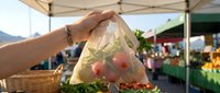Shopper holding transparent plastic bag with fresh red apples and vegetables at outdoor farmers market stand with blurred crowd and produce displays.