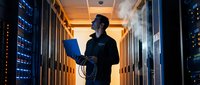 Male IT technician working with laptop in server room data center with blue LED lights and orange ambient lighting for technology infrastructure.