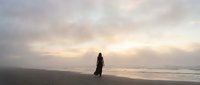 Solitary woman walking on empty beach at sunset with dramatic cloudy sky and ocean waves, peaceful coastal scene for meditation and wellness concepts.