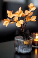 Golden metallic leaves in glass vase with black bow ribbon against dark background. Decorative branch with water drops creates elegant autumn mood.
