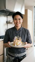 Asian male chef in dark chef's coat presenting plated gourmet dish with colorful garnishes in modern kitchen with stainless steel hood. Professional culinary portrait for restaurant, cooking, and hospitality use.