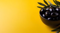 Black olives in a bowl with fresh green leaves on bright yellow background, suitable for food photography, culinary content, and recipe design.
