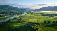 Aerial view of winding river through green valley with agricultural fields, farmhouses and forest-covered mountains under morning mist and cloudy sky.