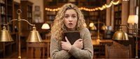 Young blonde woman with curly hair looking shocked while holding tablet in cozy library with warm lighting and vintage brass lamps for digital reaction content.