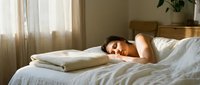 Young woman with dark hair and medium skin tone sleeping peacefully on white bedding with a pillow, in a bright bedroom with natural light from window and neutral decor, suitable for wellness, sleep, rest, and lifestyle content.