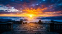 Dramatic sunset over mountains viewed from stone terrace with ornamental railings, golden hour landscape for travel and nature backgrounds.