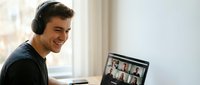 Young professional man with headphones smiling during video conference call on laptop at home office, remote work meeting with colleagues on screen.