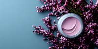 Pink moisturizing cream jar with delicate flower petals pattern on lid surrounded by fresh pink heather blooms against turquoise background, flat lay composition.