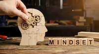 Hand placing wooden puzzle piece into head-shaped cutout with gears, representing mindset development and mental growth in workshop setting.