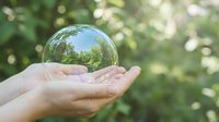 Hands holding transparent glass sphere reflecting green forest trees in natural outdoor setting symbolizing environmental protection and sustainability.