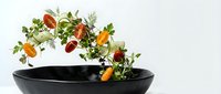 Fresh herbs and vegetables falling into black ceramic bowl on white background for healthy cooking and culinary preparation.