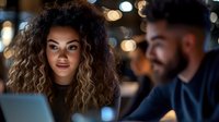 Young mixed race woman with curly hair smiling at male colleague during late night office meeting. Bokeh lights and laptop create modern workplace atmosphere.