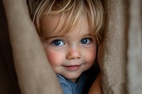 Adorable blonde toddler boy with blue eyes peeking through curtains with sweet smile. Childhood curiosity and playful hide-and-seek moment at home.