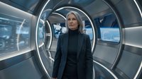 Mature woman with gray hair wearing dark blazer and turtleneck standing in futuristic corridor with glowing circular windows and blue lighting, suitable for sci-fi, technology, and corporate concepts.