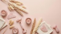 Flat lay skincare and wellness products with dried wheat, rose quartz rollers, and cosmetic jars arranged on pink background for beauty and self-care content.