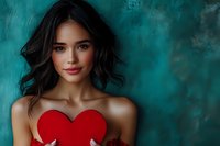Young Asian woman with dark wavy hair holding red heart shape against turquoise background. Romantic Valentine's day concept with soft dreamy atmosphere.