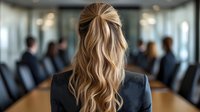 Businesswoman with long blonde wavy hair styled in half-updo, wearing dark suit jacket, viewed from behind at conference room table during corporate meeting.