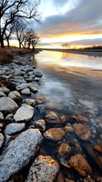 Dramatic winter sunset over river with snow-dusted rocky shore and bare trees silhouetted against golden sky, reflecting warm light on flowing water and icy stones.
