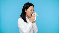 Young Asian woman with black hair wearing white long-sleeve shirt sneezing or coughing into tissue against blue background for health, illness, cold, flu, allergy concepts.