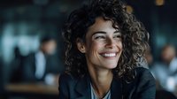 Young Mediterranean woman with curly dark hair and bright smile wearing black business suit jacket in modern office environment with blurred background. Professional corporate portrait.