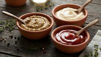Three clay bowls with different sauces and wooden spoons on rustic wooden table with herbs and spices for cooking and food preparation.