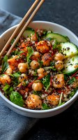 Fresh quinoa bowl with glazed chickpeas, cucumber slices, and black sesame seeds served with wooden chopsticks. Vibrant herbs and greens add color to nutritious meal.