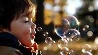 Young caucasian boy with dark hair blowing soap bubbles outdoors in autumn sunlight, capturing moment of joy and childhood wonder against bokeh background.