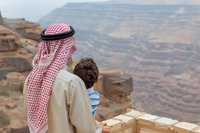 Middle Eastern man in traditional thobe and keffiyeh with child looking at dramatic desert canyon landscape from viewing point, sharing peaceful moment together.