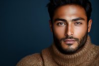 Young South Asian man with beard wearing brown turtleneck sweater against dark blue background, dramatic studio lighting highlighting facial features and confident expression.