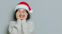 Young Asian woman wearing Santa hat and white sweater laughing with hands on face against neutral gray background, expressing holiday joy and excitement.