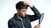 Young man with dark hair and fair complexion examining his hair loss using a handheld mirror, wearing a dark button-up shirt. Suitable for healthcare, dermatology, and wellness content.