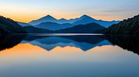 Tranquil mountain lake at dawn with perfect mirror reflection of peaks and golden sky, creating symmetrical landscape in shades of blue and orange.